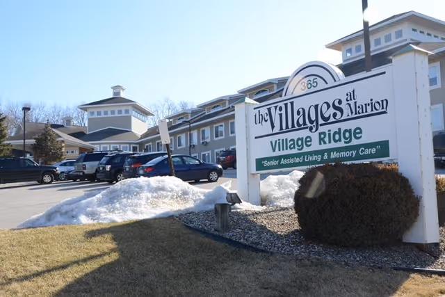 Front exterior of Village Ridge assisted living with a large sign, parked cars, and snow piles.