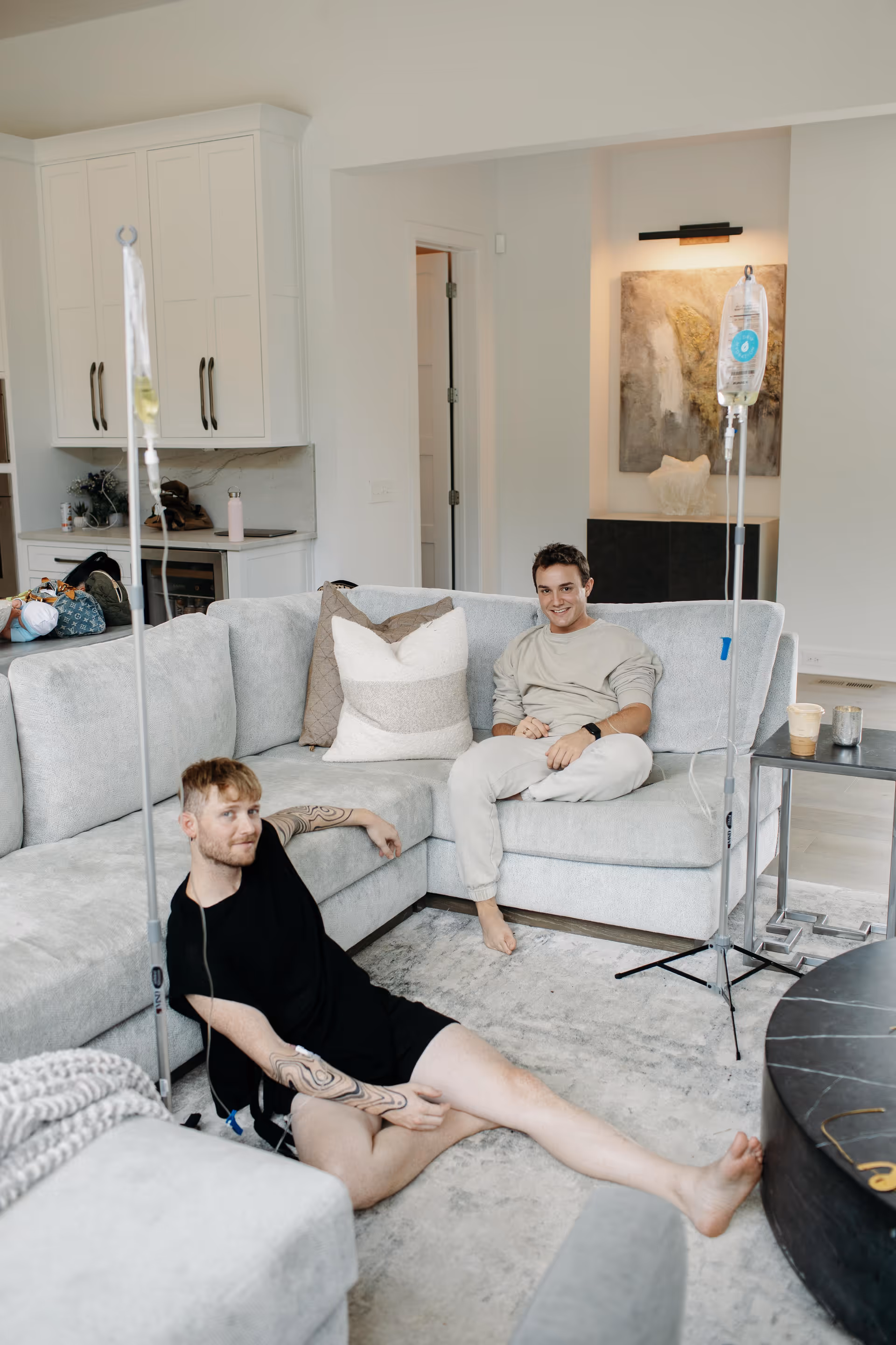 Two men sit on a gray sectional in a modern living room with IV poles.