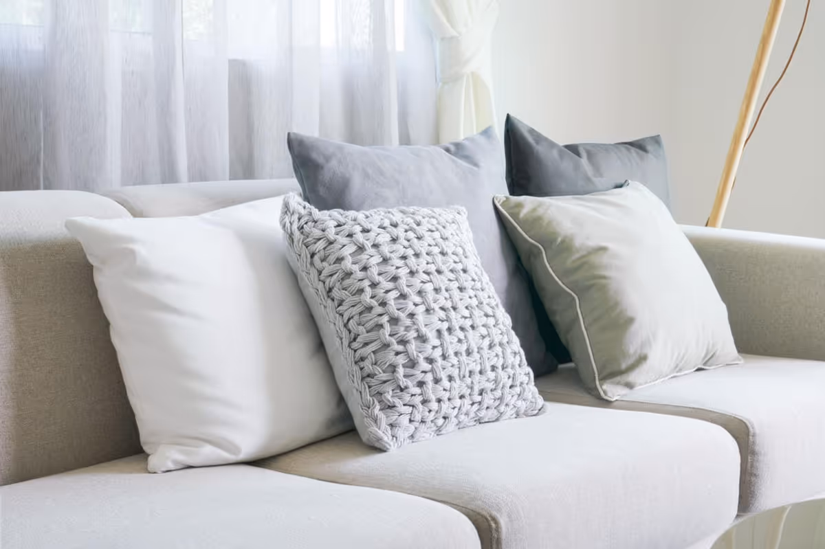A light beige sofa with four decorative pillows in shades of white, gray, and light green, placed in front of a window with sheer white curtains.