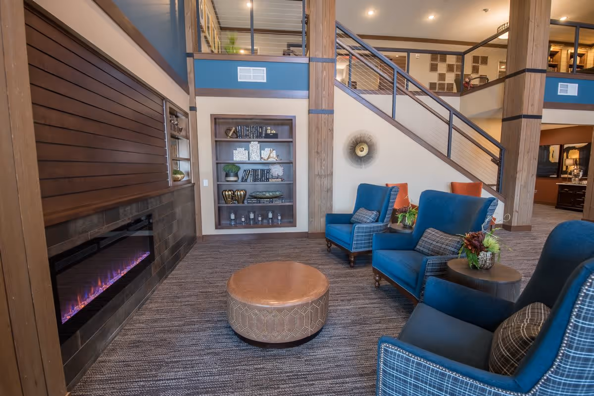 A cozy living room area with three blue armchairs arranged around a round brown ottoman. There is a modern electric fireplace on the left wall with a wooden panel above it. A built-in bookshelf with decorative items and books is set into the wall. A staircase with metal railings leads to an upper floor, and the space is warmly lit with recessed ceiling lights.