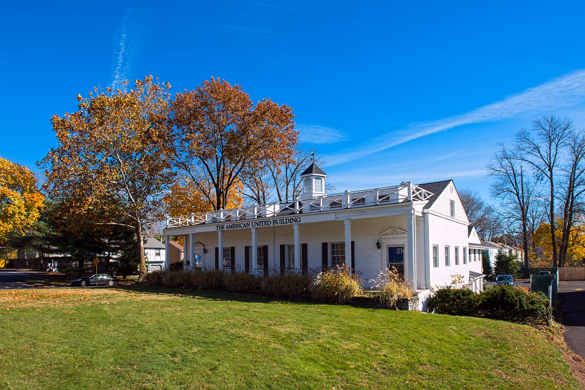 White building with a wraparound porch sits on green lawn under autumn trees and blue sky.