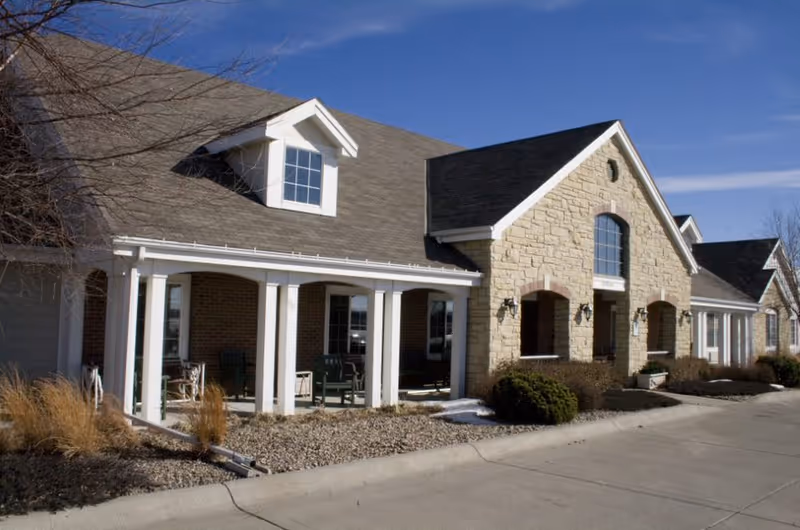 Exterior view of a single-story assisted living facility building with a stone and brick facade, white columns supporting a covered porch, several windows, and a clear blue sky above.