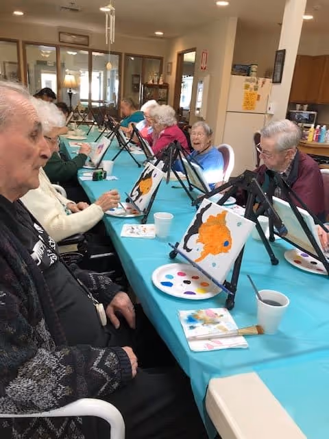 A group of elderly individuals seated around a long table covered with a turquoise tablecloth, engaged in a painting activity. Each person has a small easel with a canvas featuring a partially painted orange fish, along with paint palettes and brushes. The room appears to be a common area with bright lighting and a kitchen visible in the background.