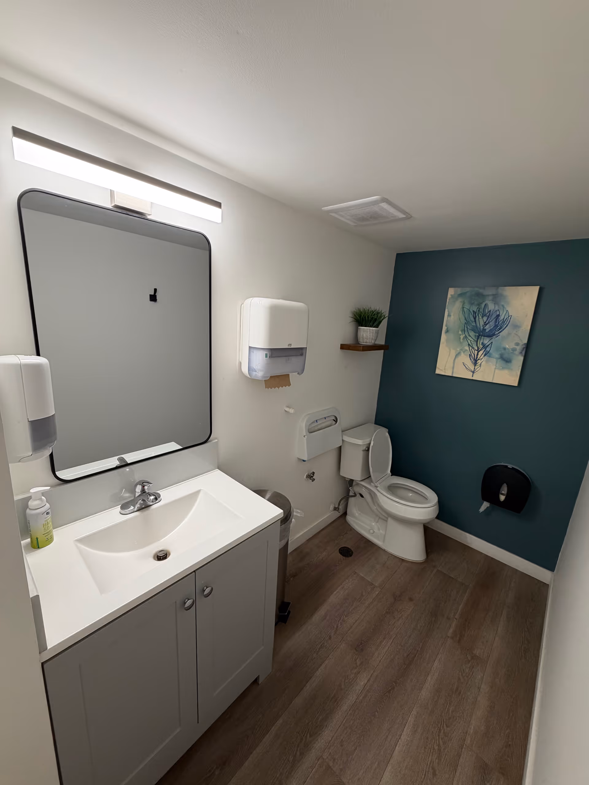 Small modern bathroom with sink, mirror, toilet, and teal accent wall.