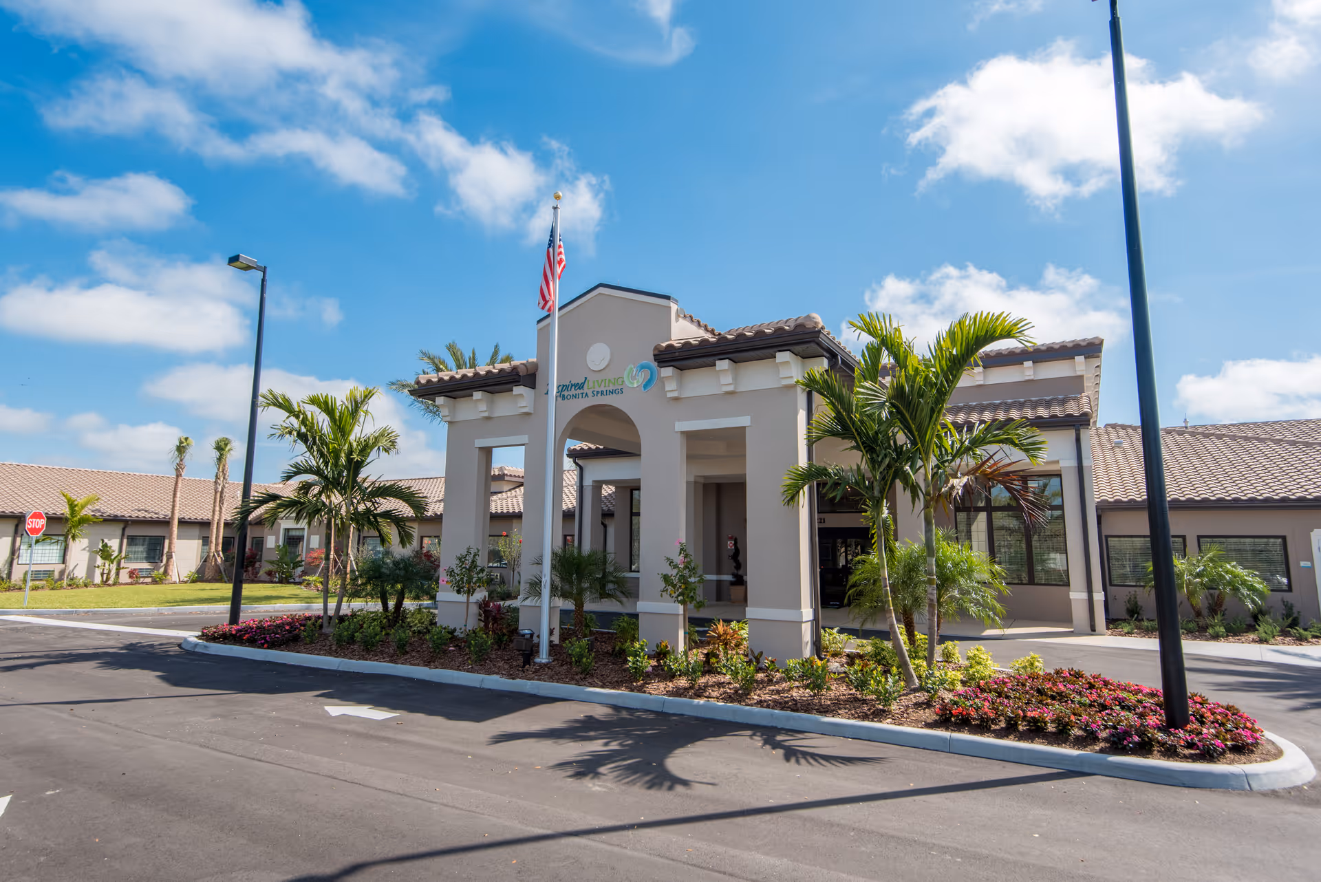 Exterior view of Inspired Living Bonita Springs facility with a beige building featuring a covered entrance, palm trees, landscaped flower beds, an American flag on a flagpole, and a clear blue sky with some clouds.
