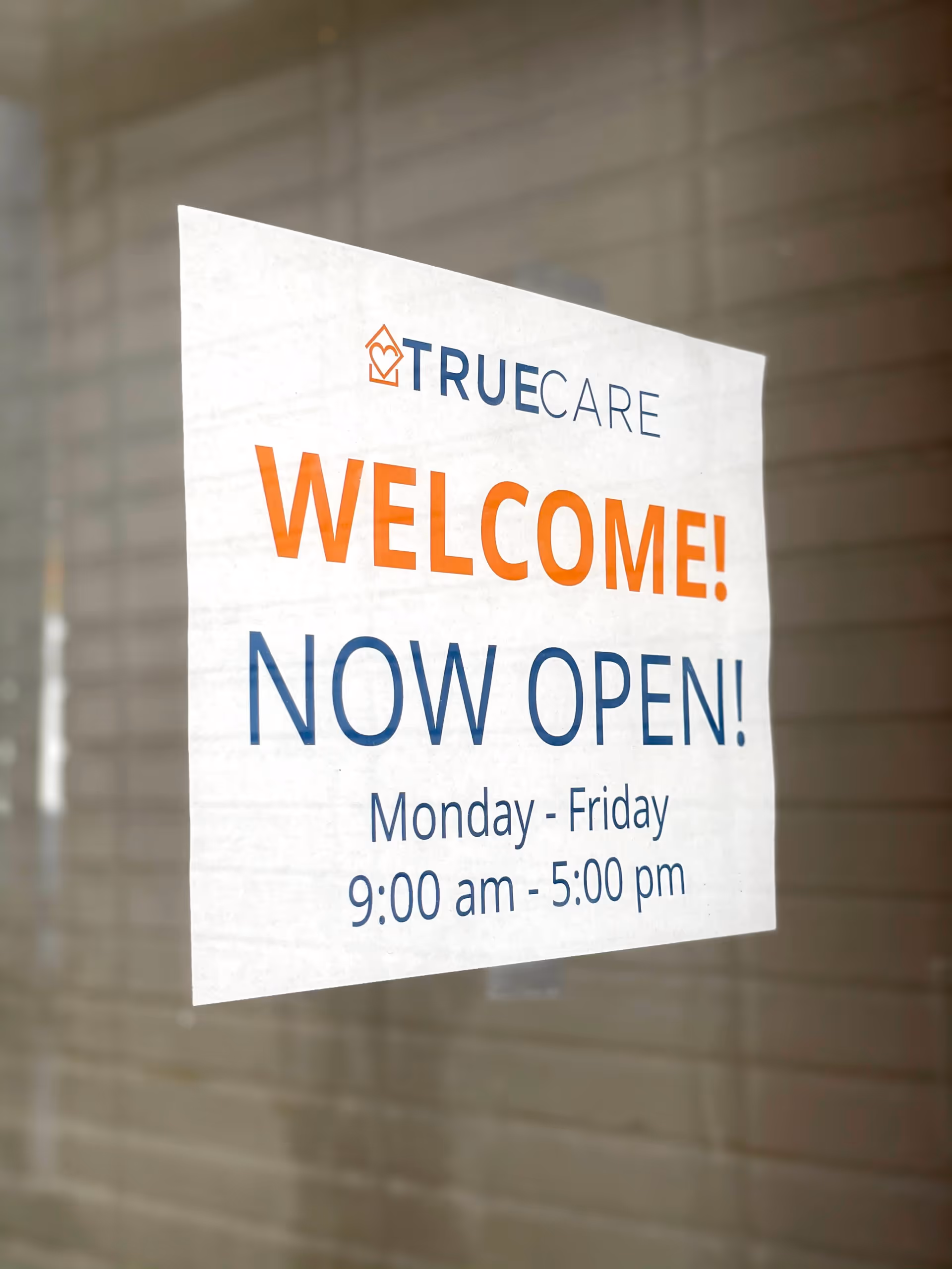 White sign on glass window announces TrueCare Now Open with hours outside a building.
