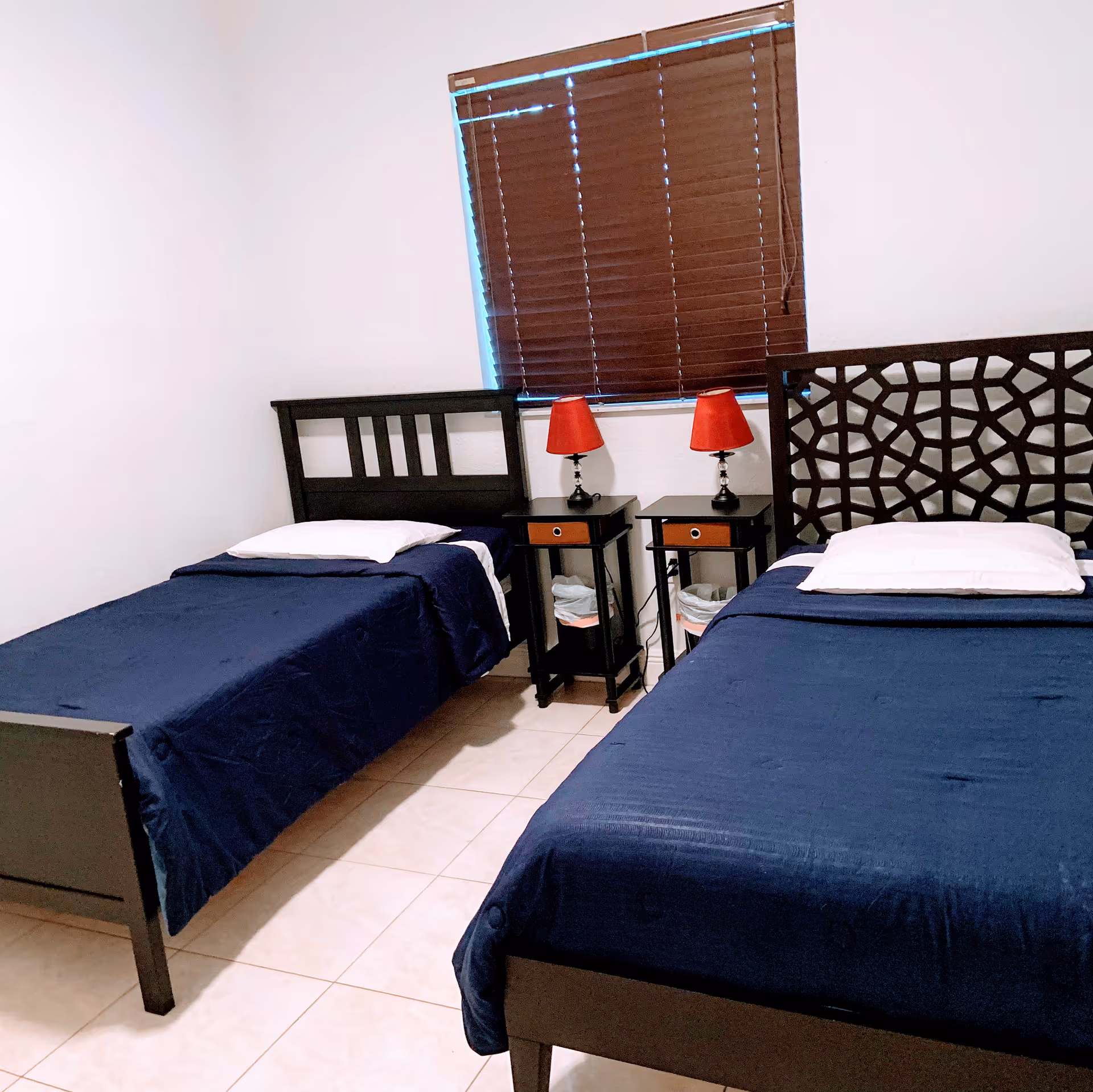 A bedroom with two single beds covered in dark blue bedspreads and white pillows. Between the beds are two small black nightstands, each with a red lampshade on a lamp. The window behind the nightstands has closed brown blinds. The floor is tiled in a light color.