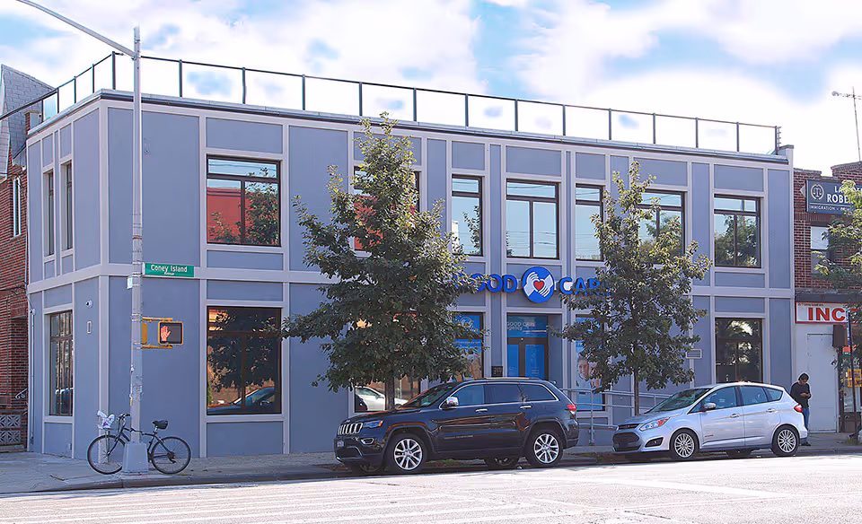 Blue-gray two-story building with Good Care Agency sign, street corner, trees, and parked cars.