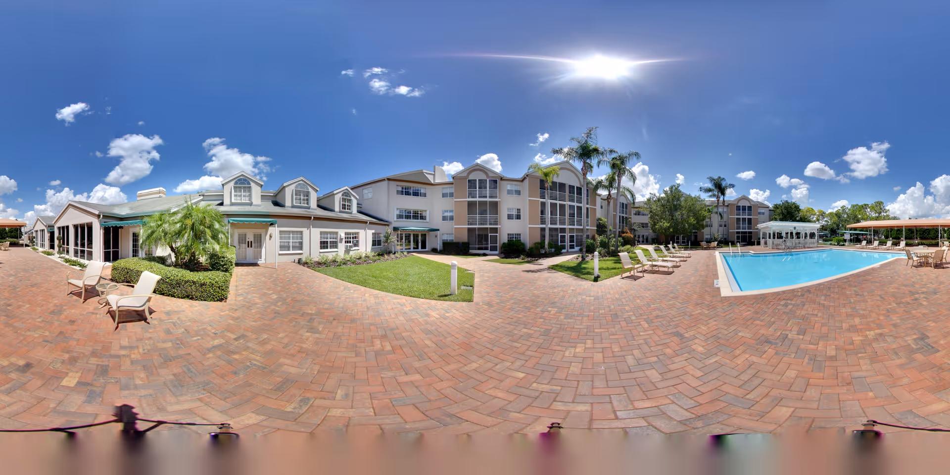 Outdoor view of Coral Oaks senior living facility showing a large swimming pool surrounded by lounge chairs, a brick-paved patio area, palm trees, and multi-story residential buildings under a bright sunny sky with scattered clouds.