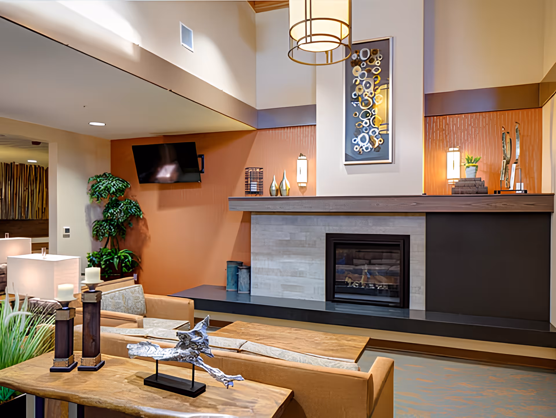 A cozy living room area in Avamere Transitional Care of Puget Sound featuring a modern fireplace with a wooden mantel, decorative vases, and a framed abstract artwork above it. The room includes comfortable beige armchairs, a wooden coffee table with decorative items, a flat-screen TV mounted on an orange accent wall, and a tall green plant in the corner. Soft lighting from lamps and a hanging light fixture create a warm ambiance.