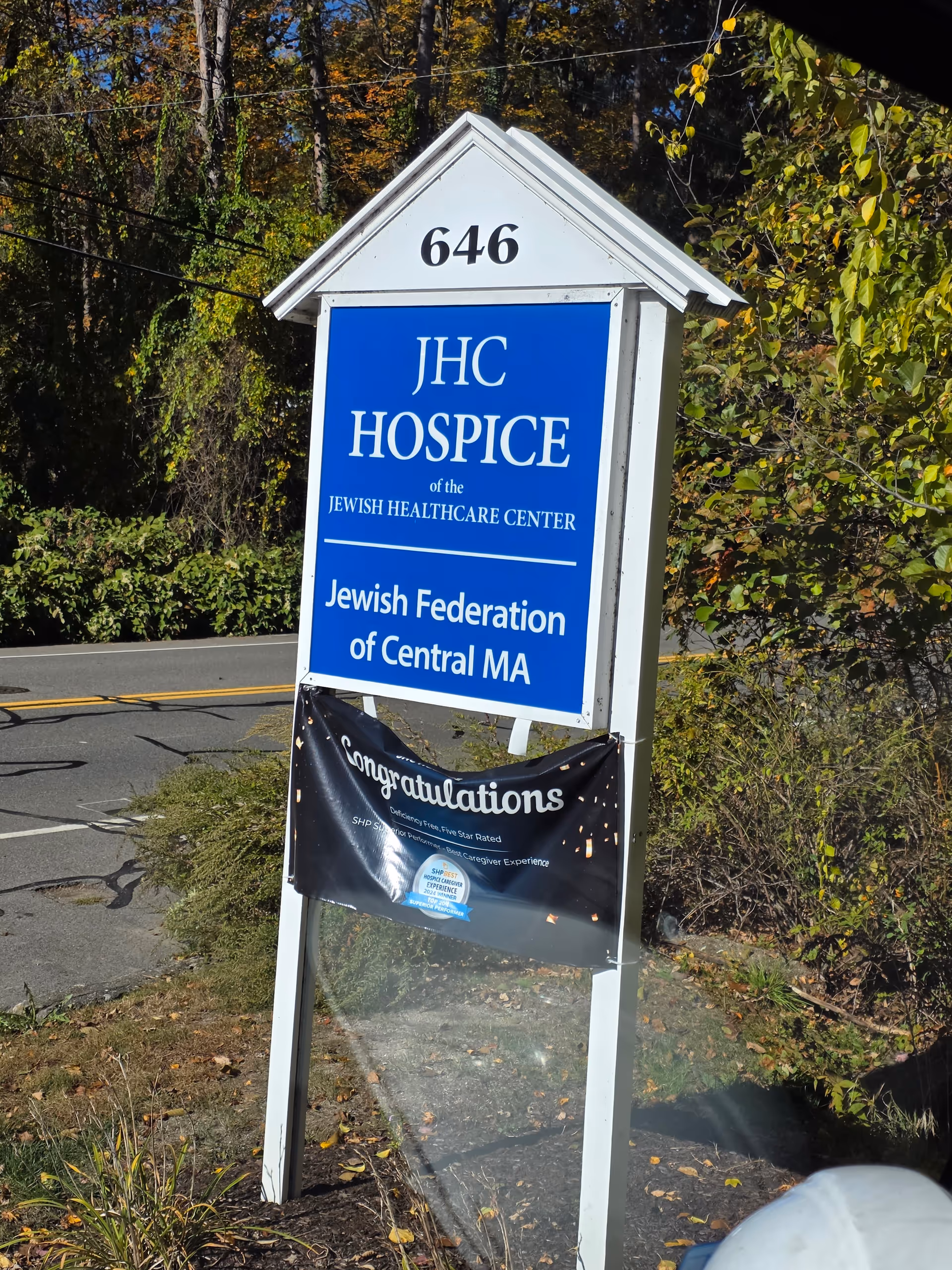 Blue JHC Hospice sign on a post with trees and a 'Congratulations' banner.