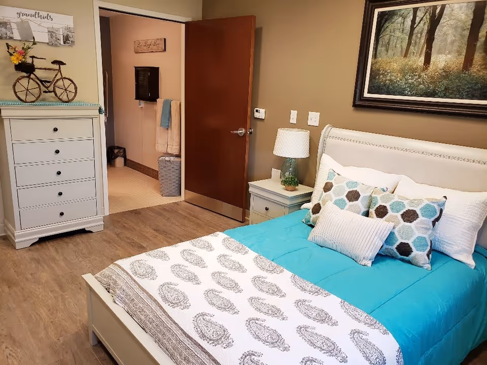 Cozy furnished bedroom with a teal comforter, patterned pillows, nightstand and dresser, and an open door leading to a bathroom.