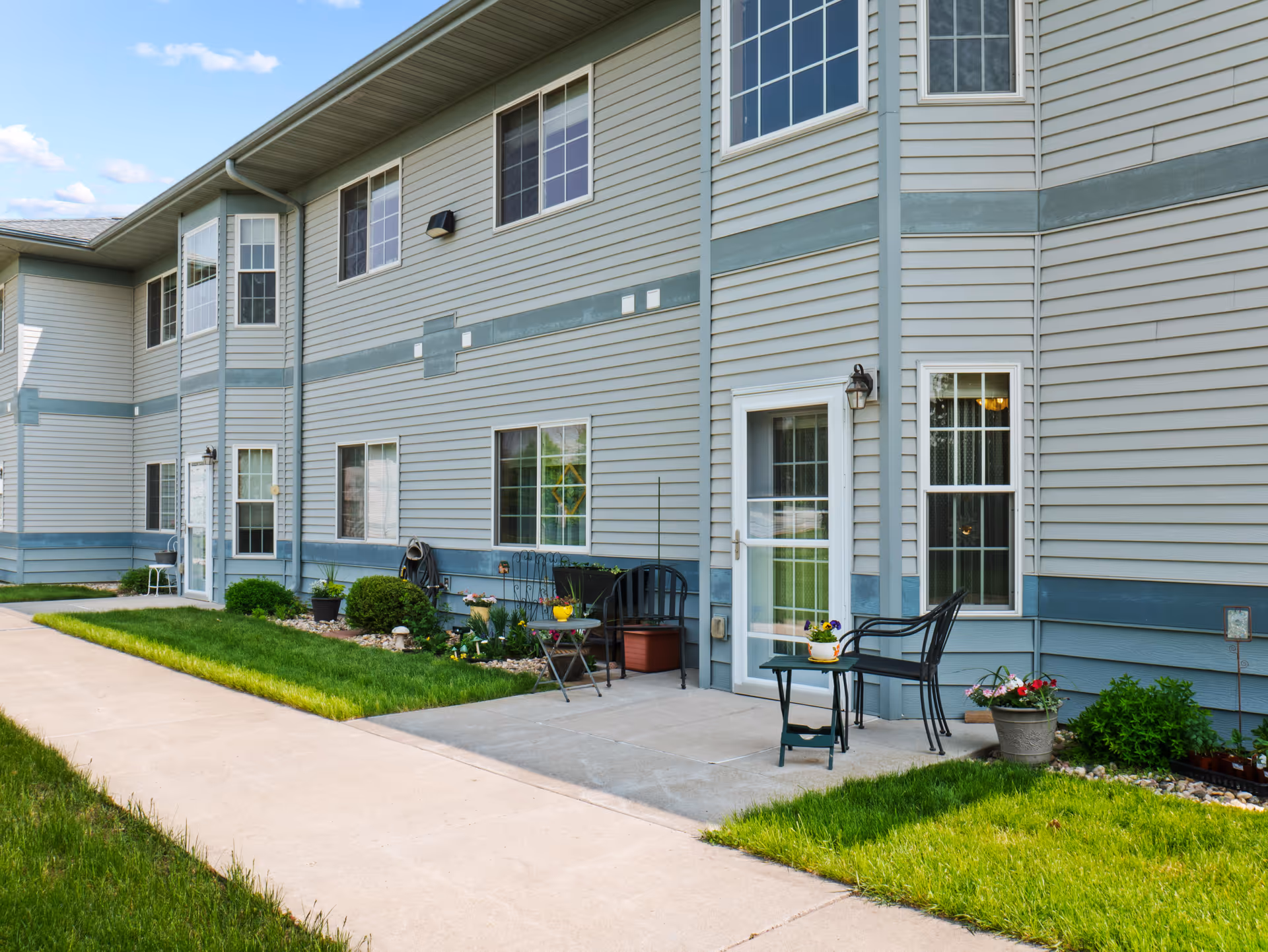 Exterior view of a two-story residential building with light gray siding and blue trim. There is a concrete walkway and small patio areas with chairs, tables, and potted plants. The lawn is green and well-maintained under a partly cloudy sky.