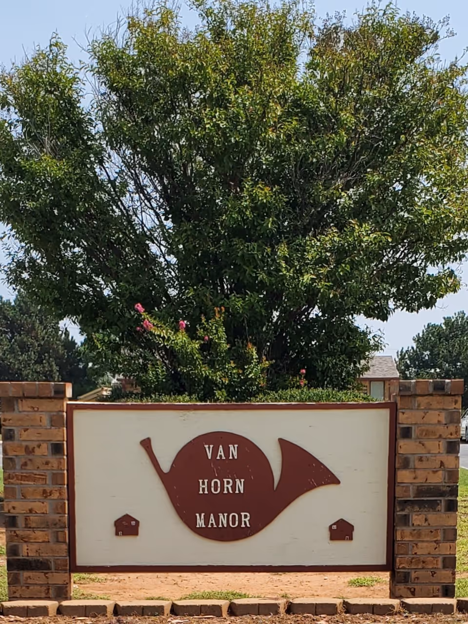 Exterior sign for Van Horn Manor with brick pillars and a large tree behind.