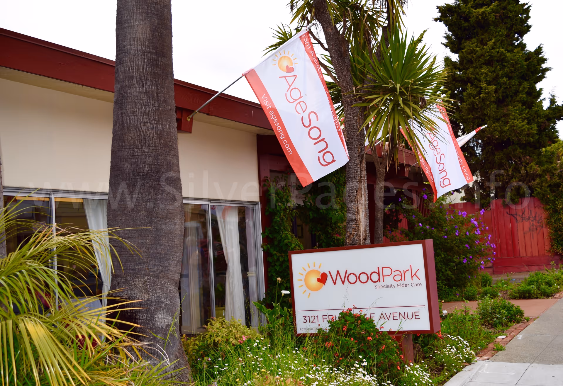 Exterior of Elder Ashram senior care facility with WoodPark sign, banners, and landscaping near entrance.
