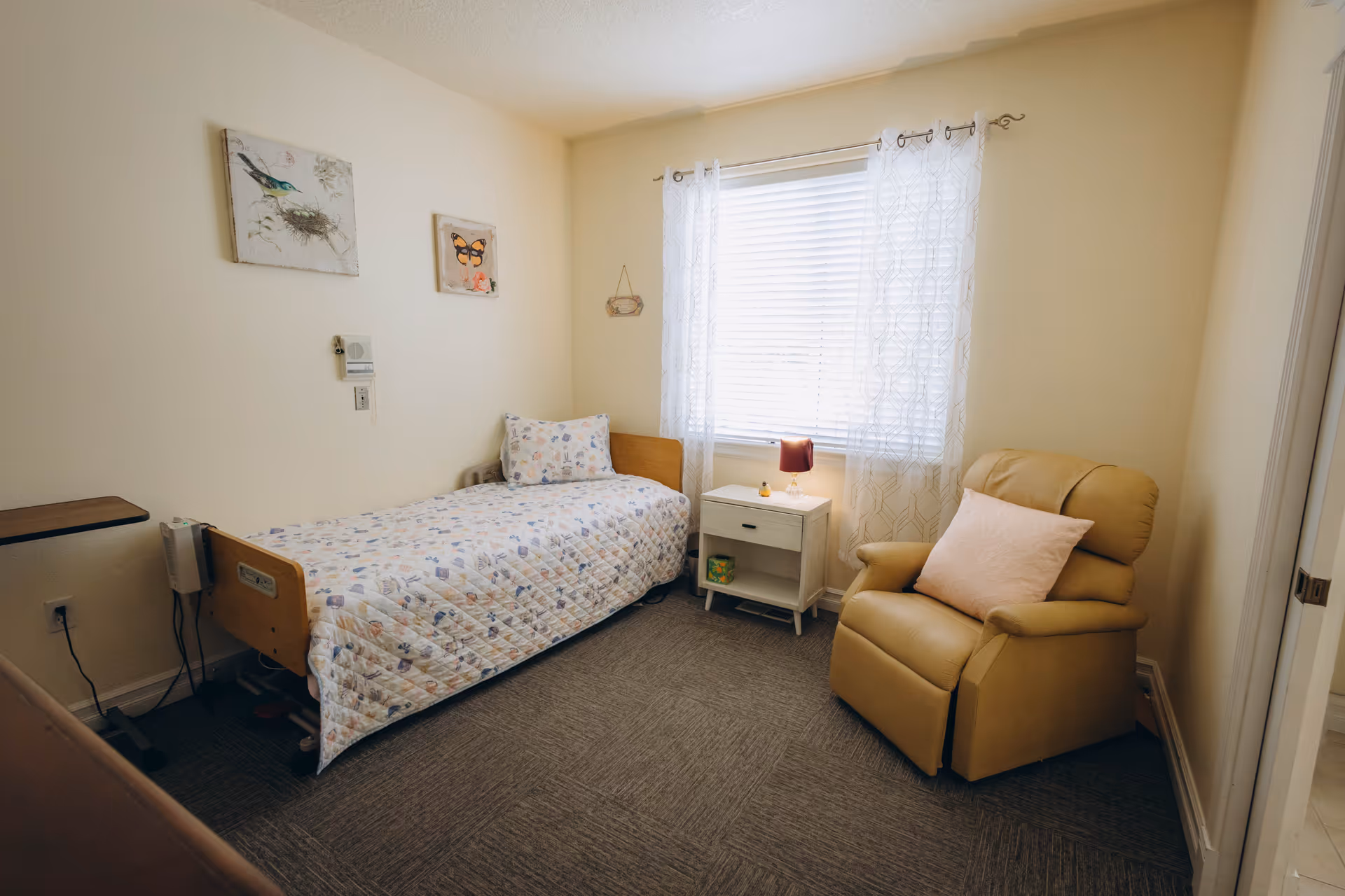 Small furnished bedroom with single bed, nightstand, recliner, and window with sheer curtains