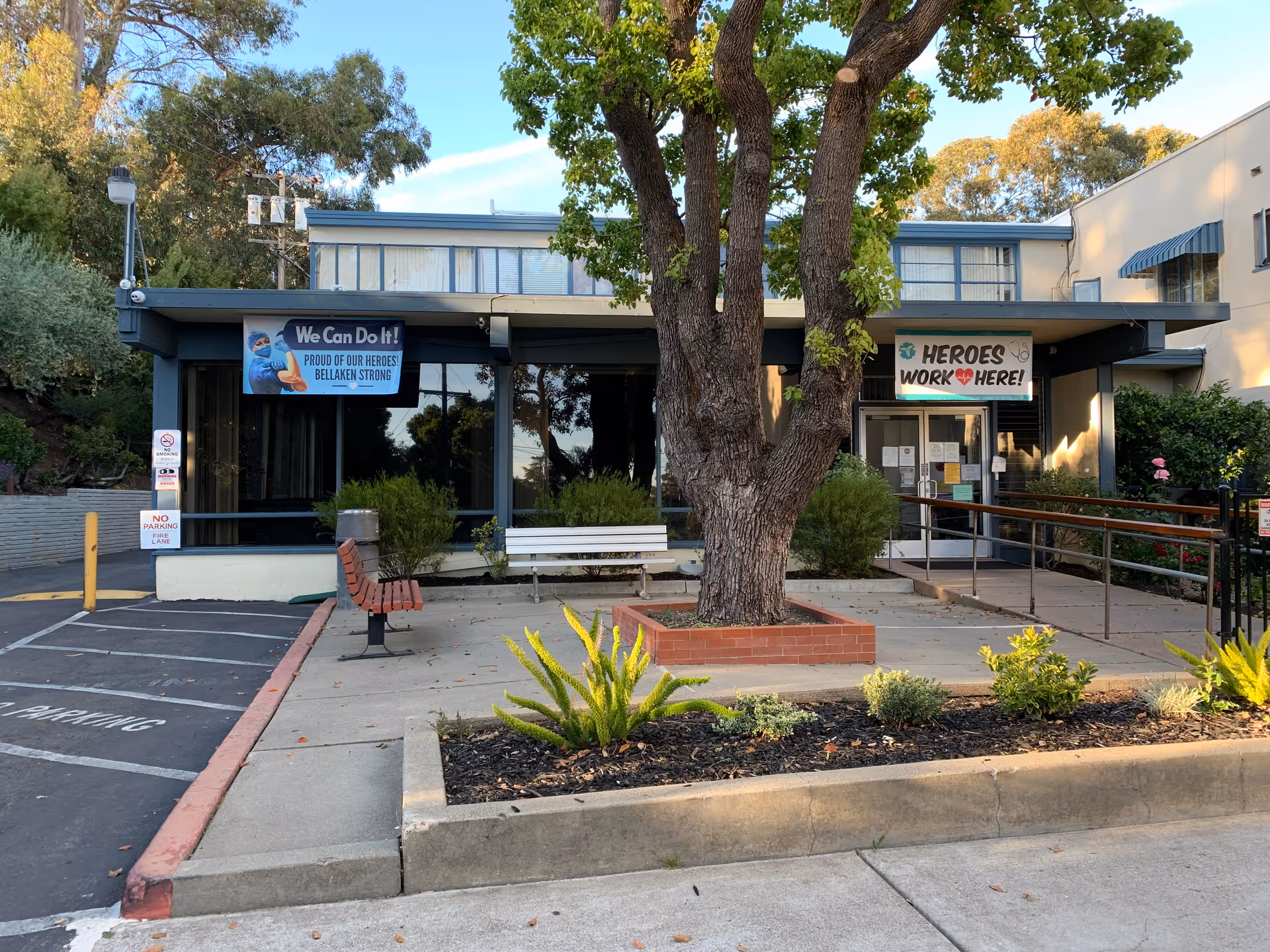 Front entrance of Bellaken Garden senior living facility with a large tree, benches, planters, a wheelchair ramp and banners reading 'We Can Do It!' and 'Heroes Work Here!'