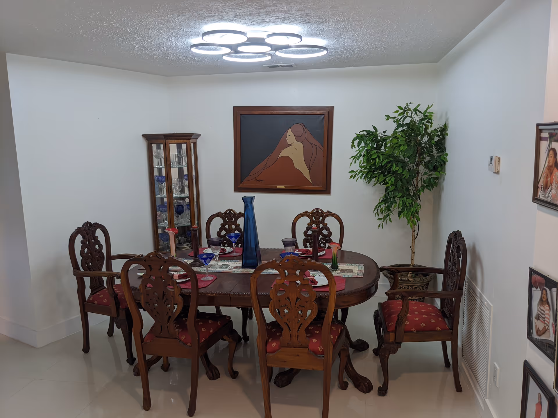 Round wooden dining table with six ornate chairs, glass cabinet, painting, and a tall plant.