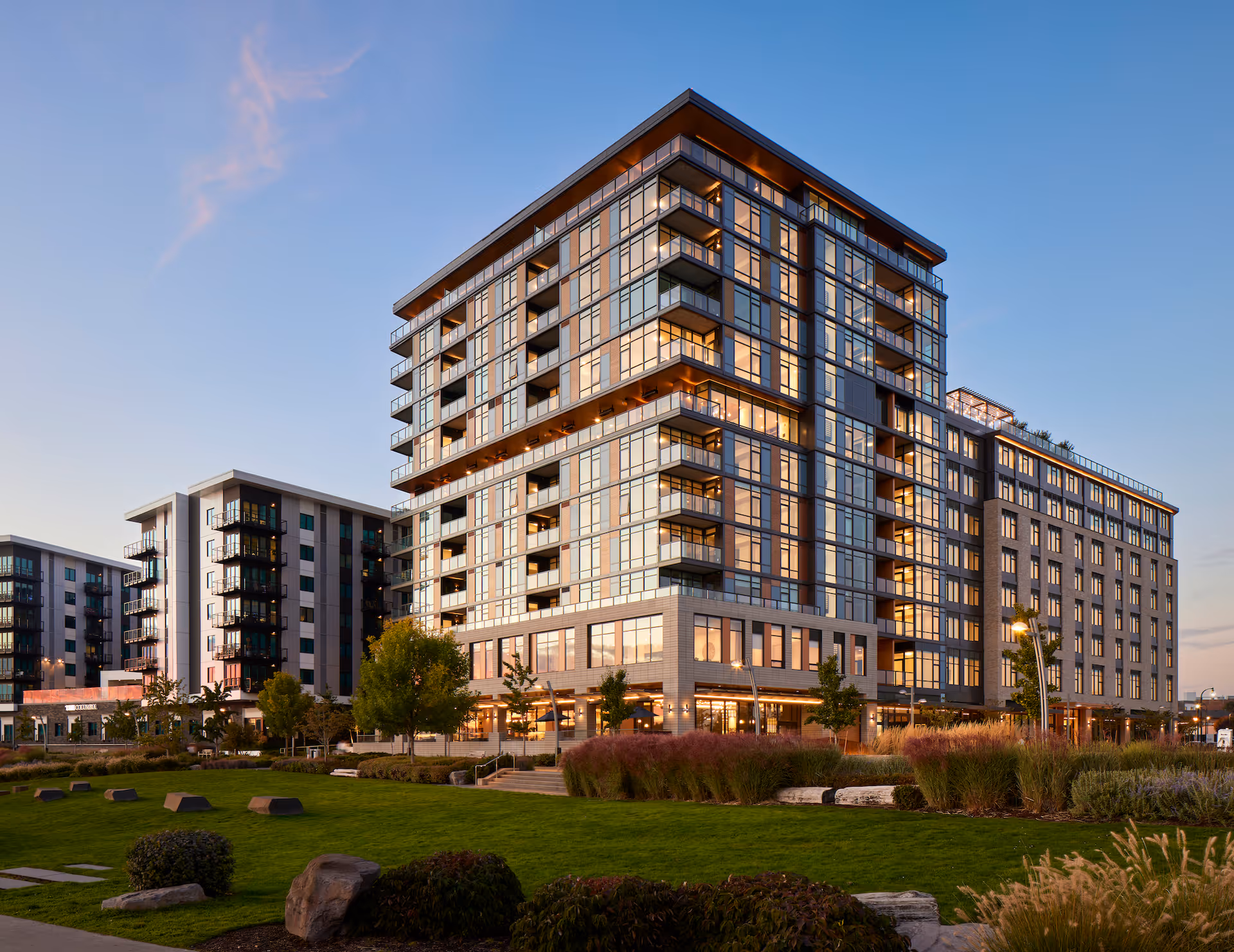 Modern multi-story residential building with large windows and balconies, surrounded by landscaped green lawn and trees under a clear evening sky.