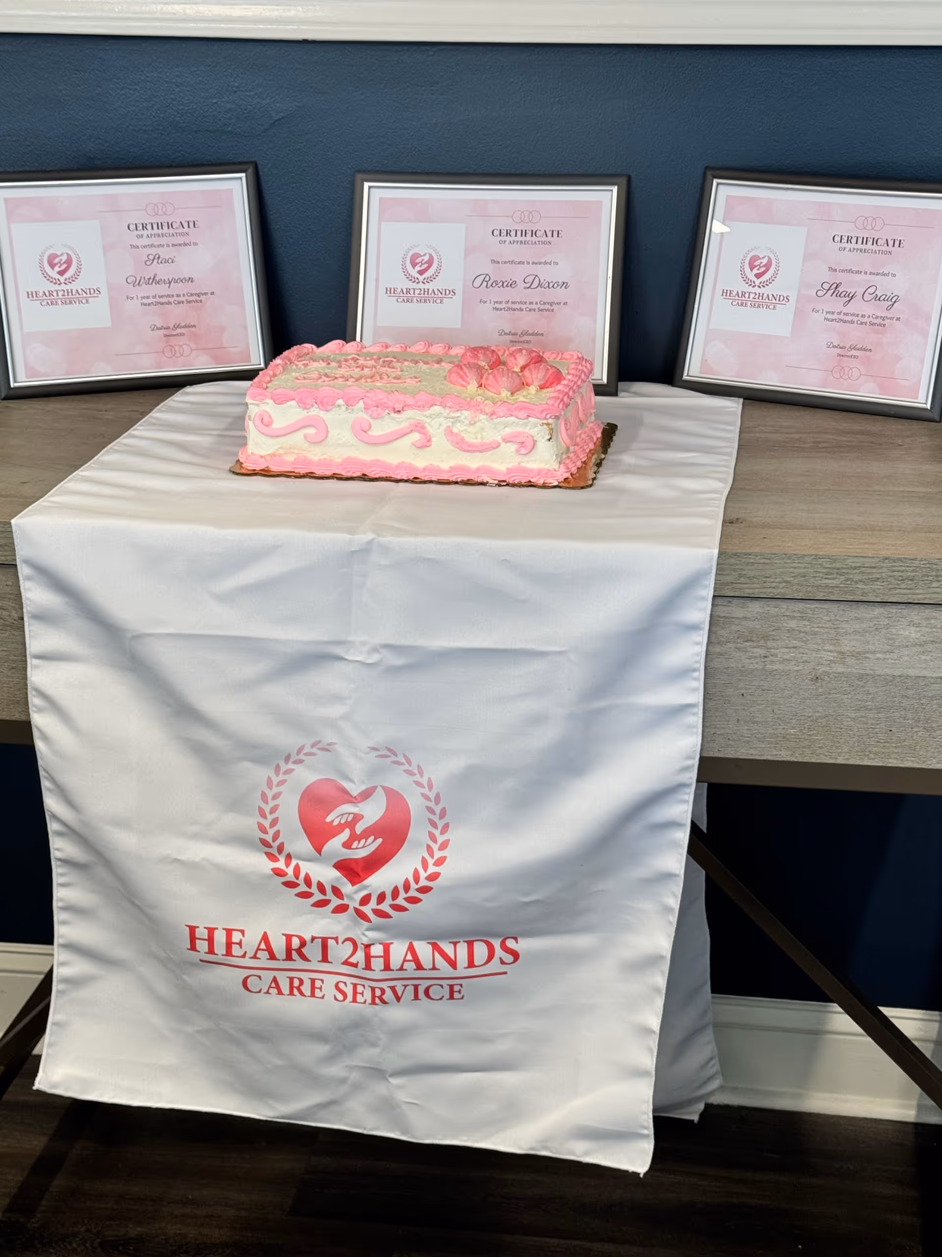 Pink decorated cake sits on a table, with Heart2Hands certificates displayed behind.