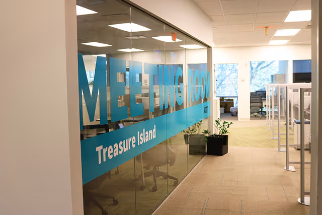Office hallway shows a glass wall meeting room sign reading Meeting Room Treasure Island and plants.