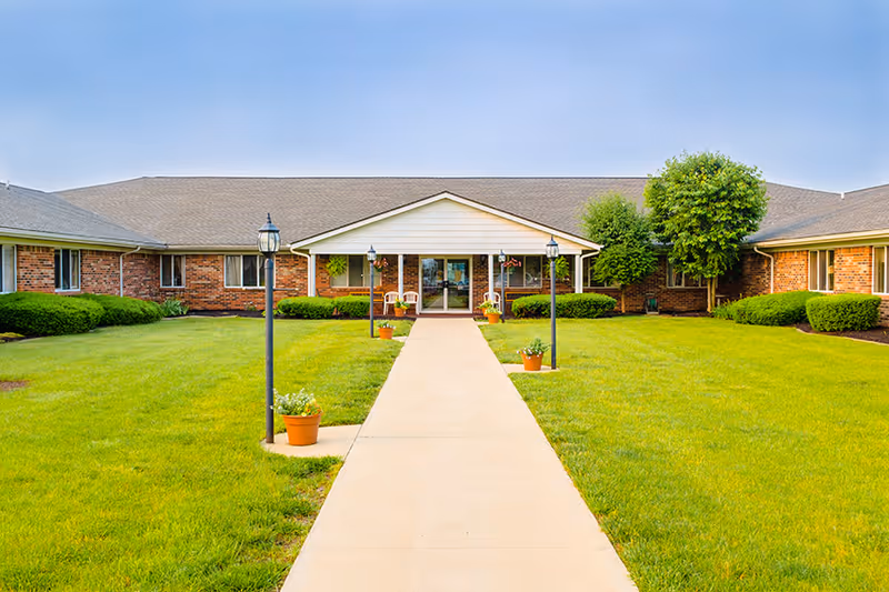 Front exterior view of Clinton House Rehabilitation and Healthcare Center showing a single-story brick building with a central entrance covered by a white gabled roof. A concrete walkway leads to the entrance, flanked by lamp posts and potted plants. The surrounding lawn is green and well-maintained with bushes and trees near the building.