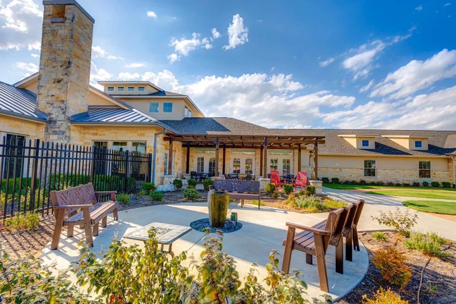 Outdoor courtyard with benches, a small fountain and a pergola in front of a stone-clad senior living building under a blue sky.