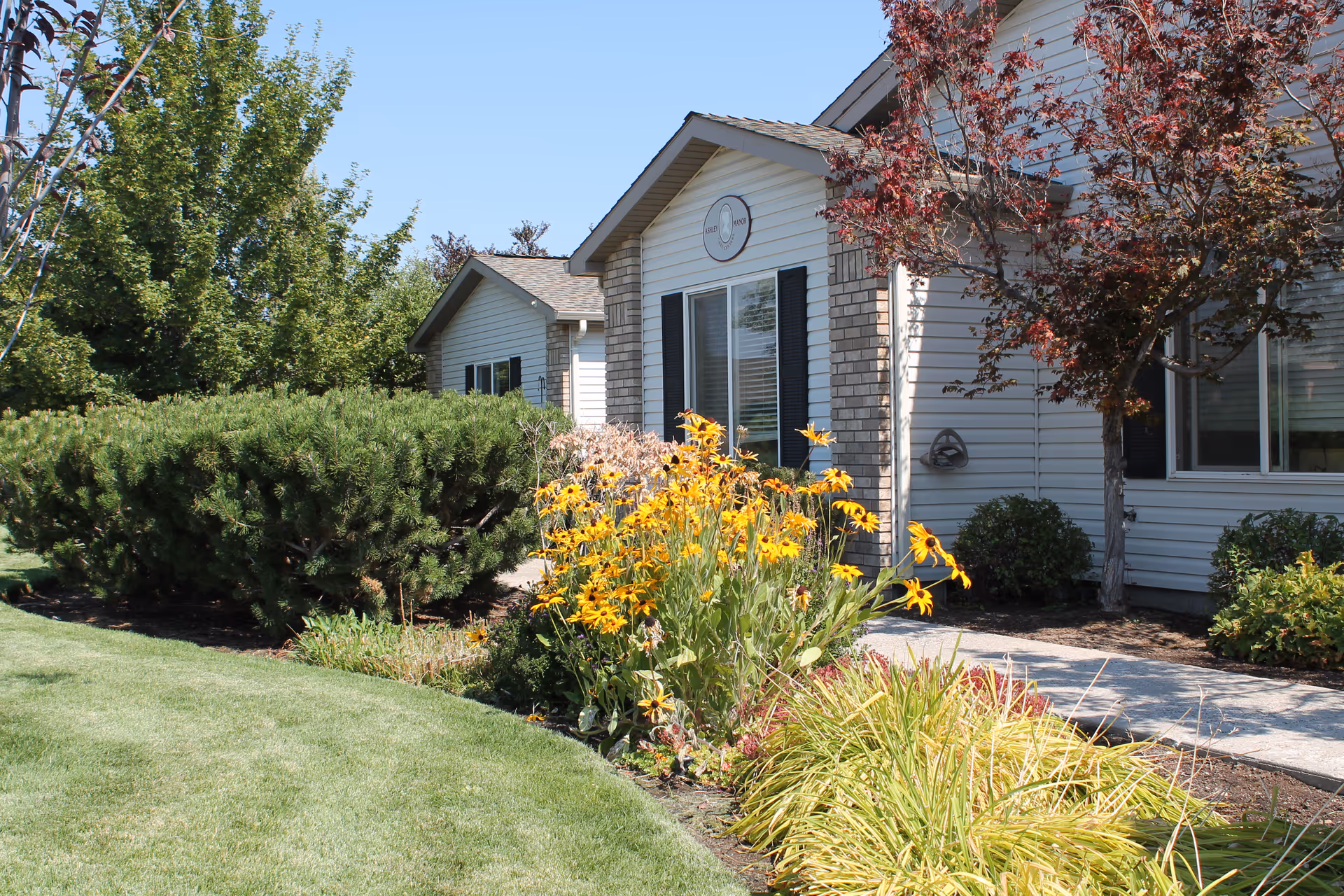 Exterior view of a single-story building with white siding and brick accents, surrounded by a well-maintained garden featuring yellow flowers, green shrubs, and trees under a clear blue sky.