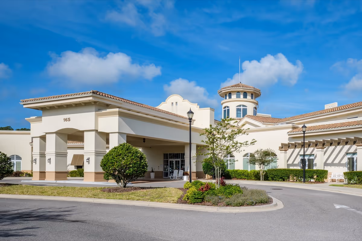 Front entrance of a Mediterranean-style senior living facility with a porte-cochère, landscaped circular driveway, and a round tower under a blue sky.