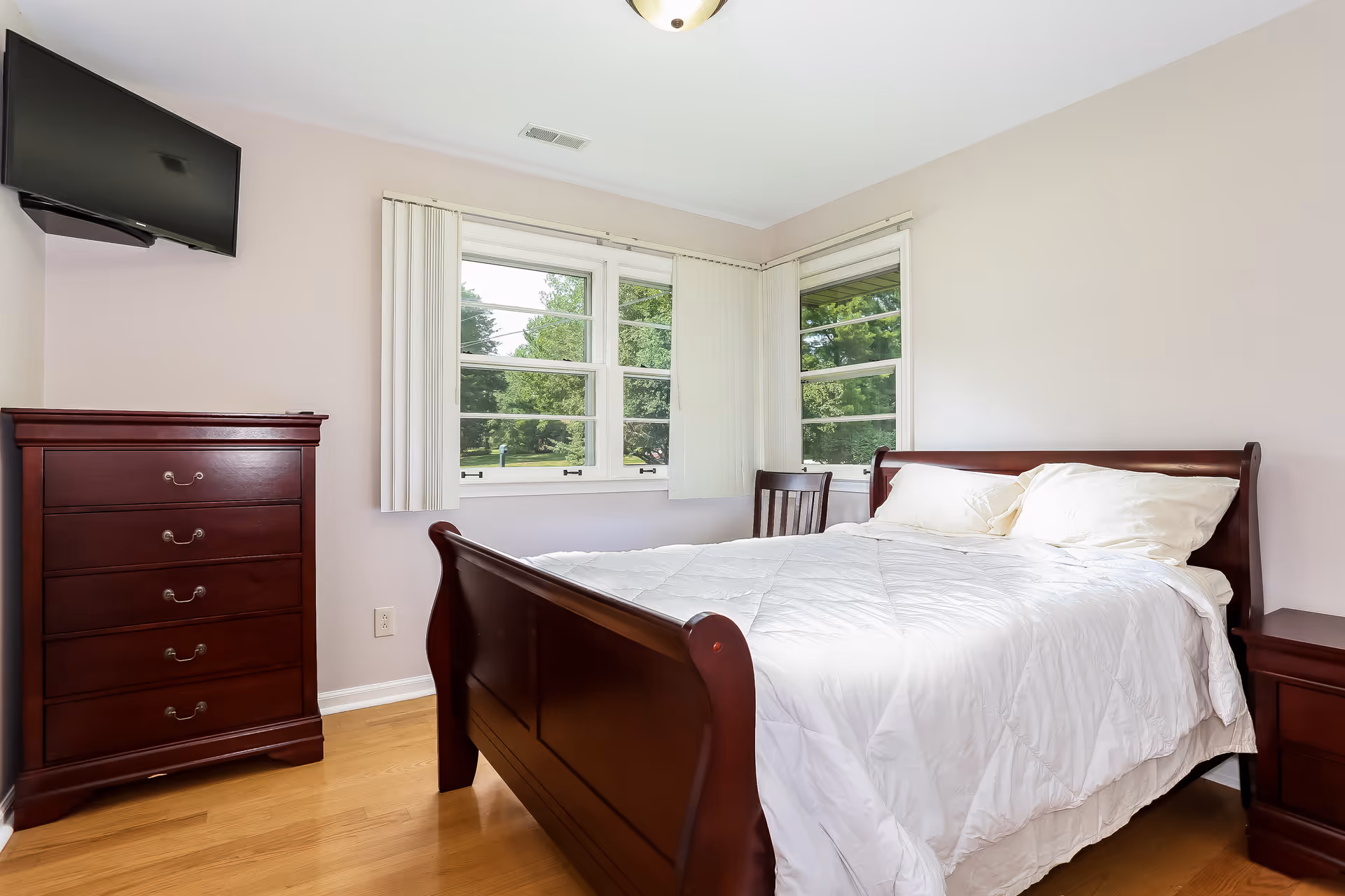 A cozy bedroom features a wooden bed, dresser, and two windows overlooking greenery.