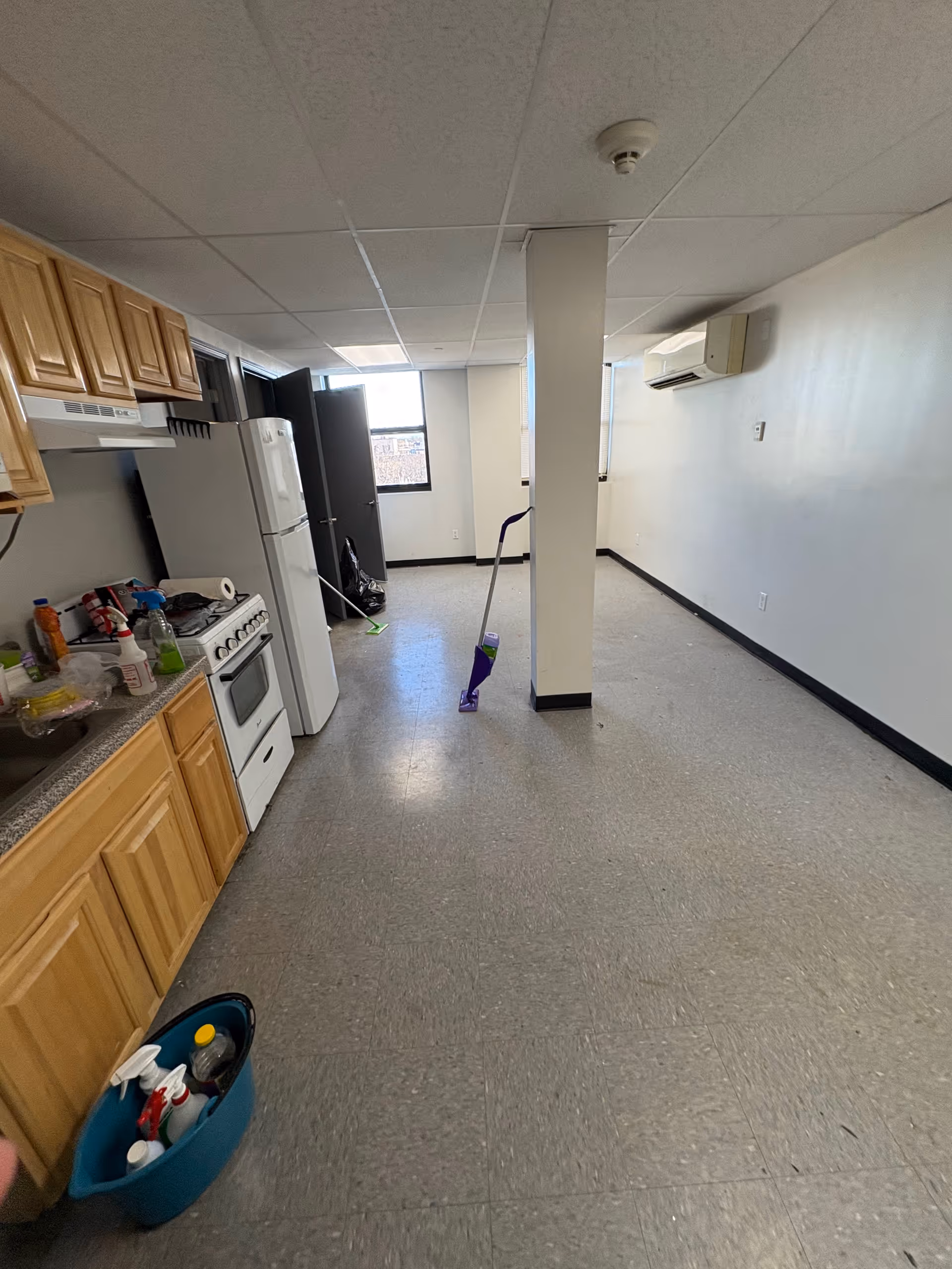 Long kitchen area with wooden cabinets, fridge, stove, mop, and cleaning supplies in a senior facility.