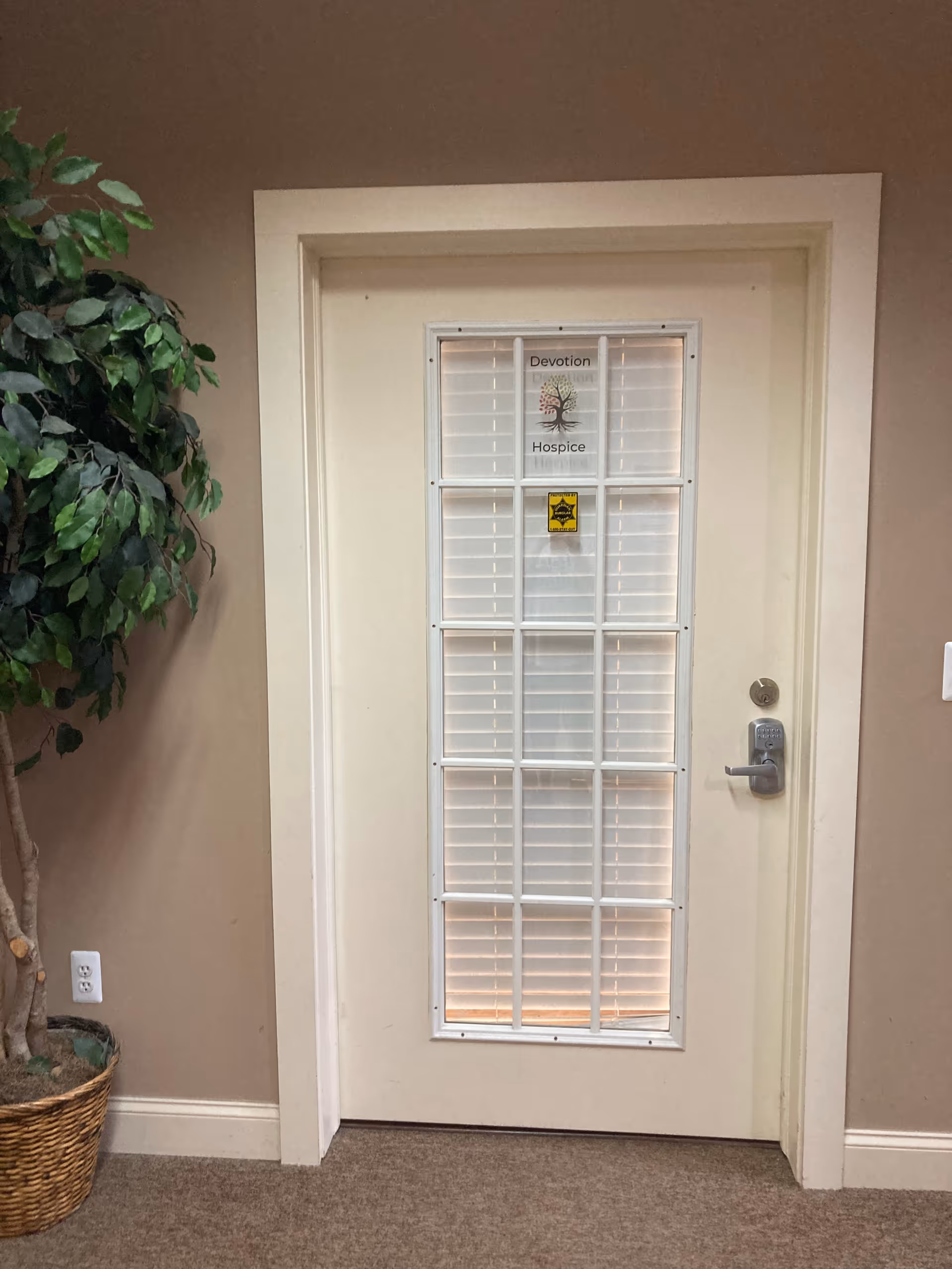 Cream-colored interior door with glass panes and blinds labeled Devotion Hospice, beside a potted plant.