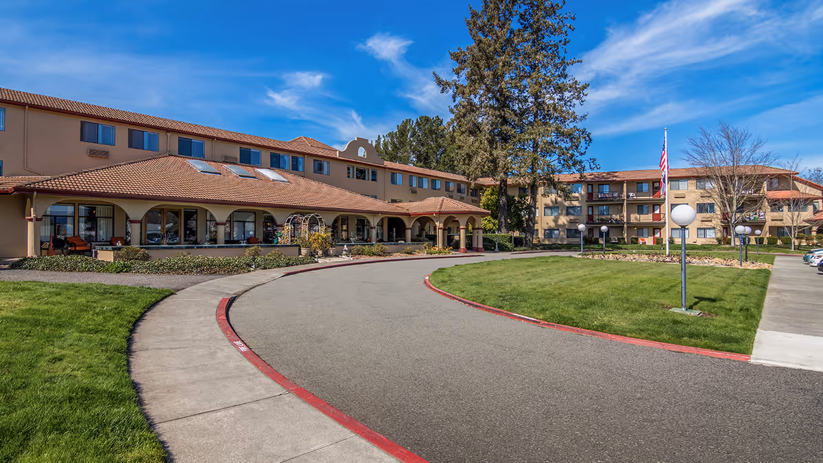 Front exterior of a multi-story senior living building with a curved driveway, landscaped lawn, and arched entrance.