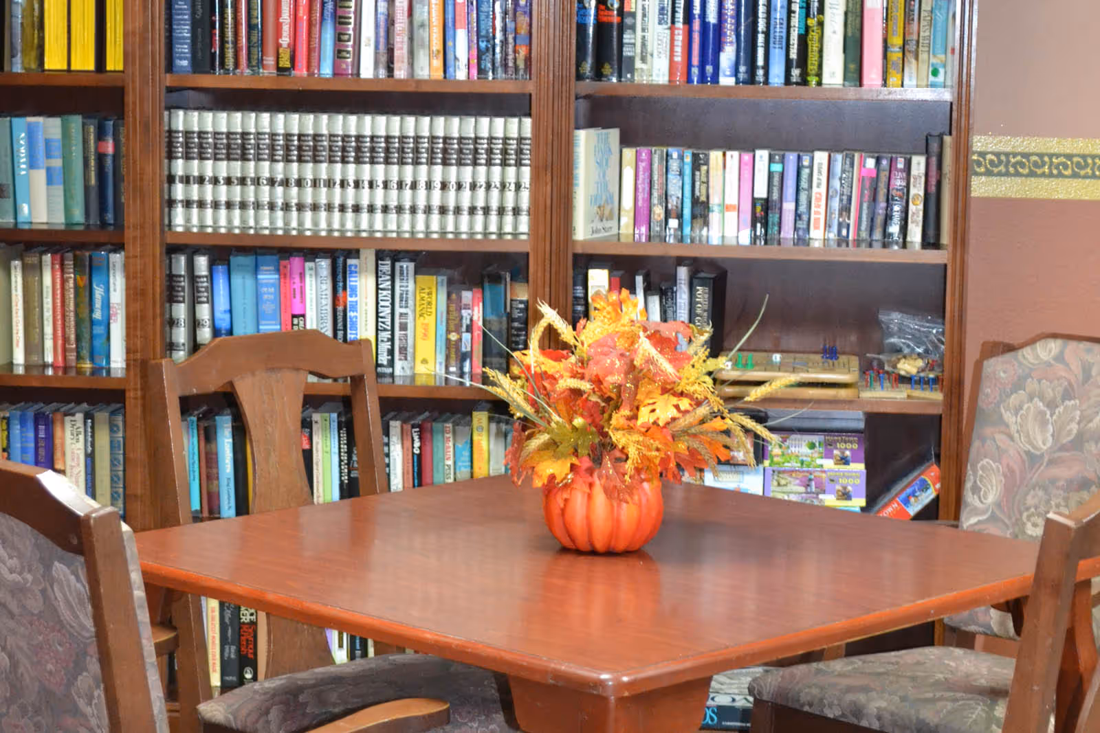 A wooden table with a fall-themed floral centerpiece in a pumpkin-shaped vase, surrounded by wooden chairs with floral upholstery. Behind the table is a large bookshelf filled with various books and board games.