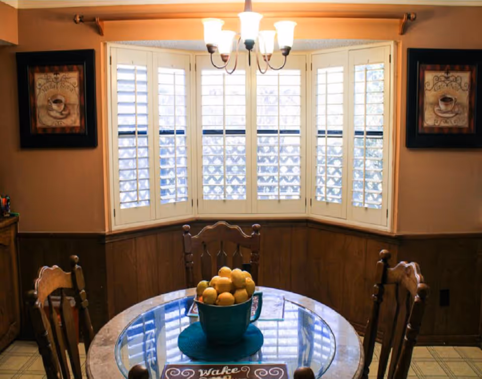 A cozy dining room with a round glass table, wooden chairs, and a bay window.