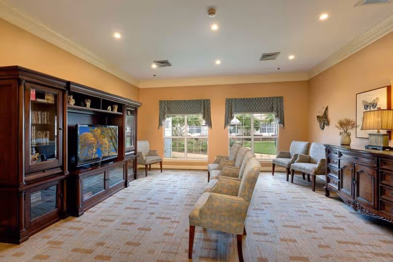 Bright common sitting room with upholstered chairs arranged facing a large wooden entertainment center and windows overlooking a courtyard.