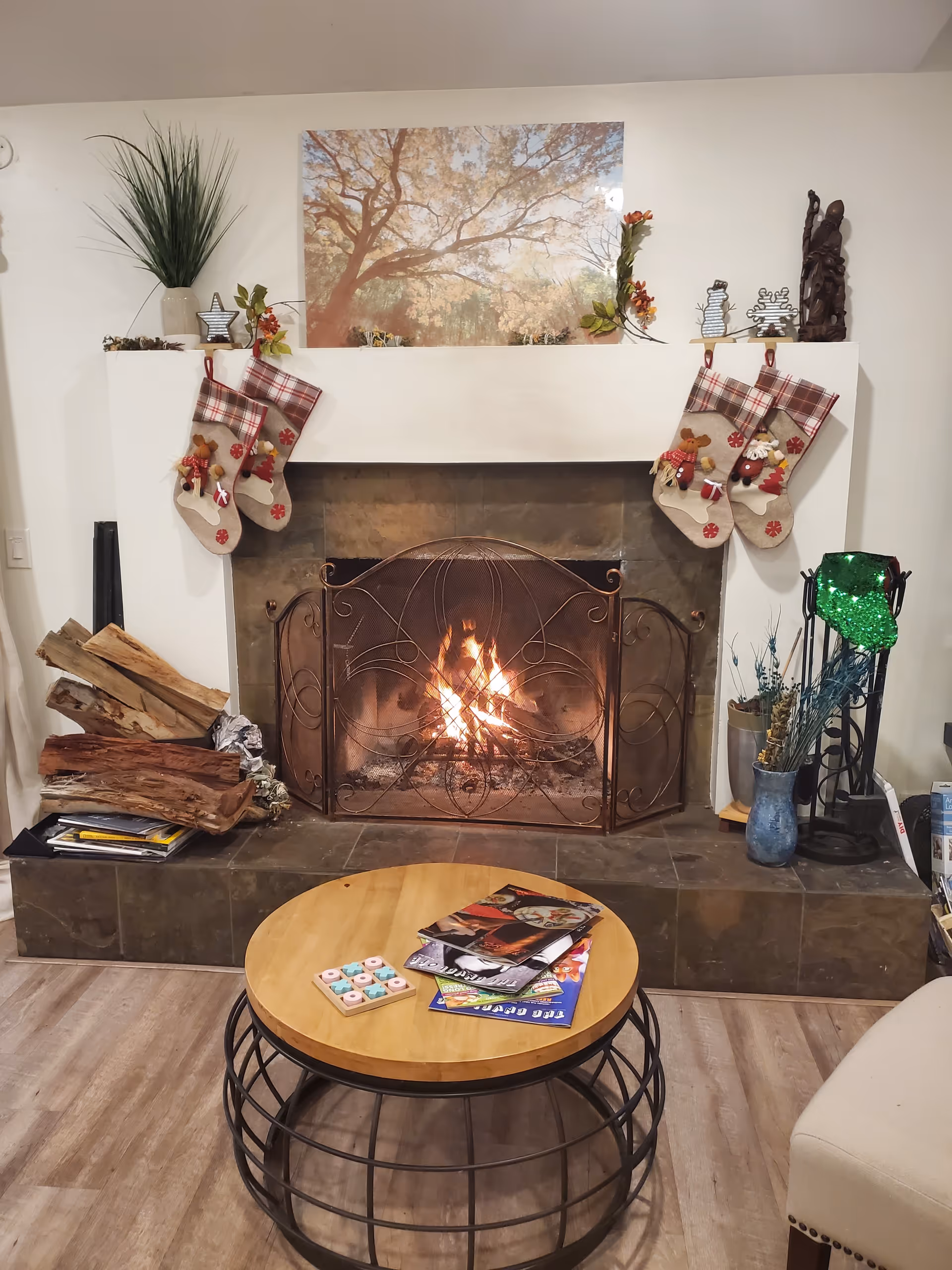 A cozy living room scene featuring a lit fireplace with a decorative metal screen. Four Christmas stockings with plaid cuffs and festive decorations hang from the white mantel. On the mantel, there are small decorative items and a large framed picture of a tree with autumn leaves. To the left of the fireplace, there is a stack of firewood and some magazines or books. To the right, there is a blue vase with dried flowers and a green decorative item on a stand. In front of the fireplace is a round wooden coffee table with a black metal frame, holding magazines and a small game. Part of a beige upholstered chair is visible on the right side.