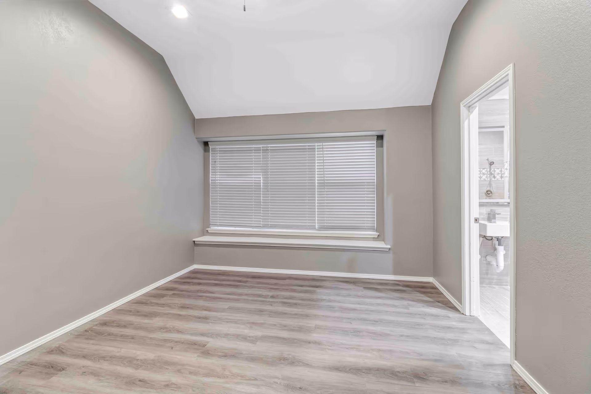 Empty light-gray bedroom with wood-look flooring, a large window with blinds and a small window seat, and a doorway leading to a bathroom.