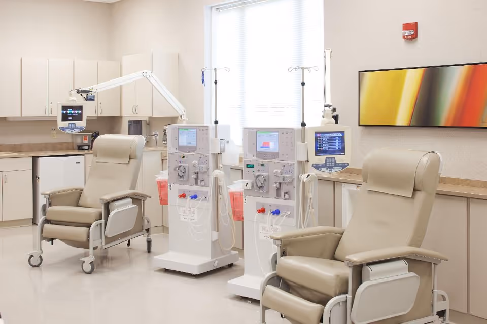 A clean medical treatment room with two beige recliner chairs on wheels, two dialysis machines with attached IV poles, white cabinets and countertops, a small refrigerator, and a colorful abstract painting on the wall.