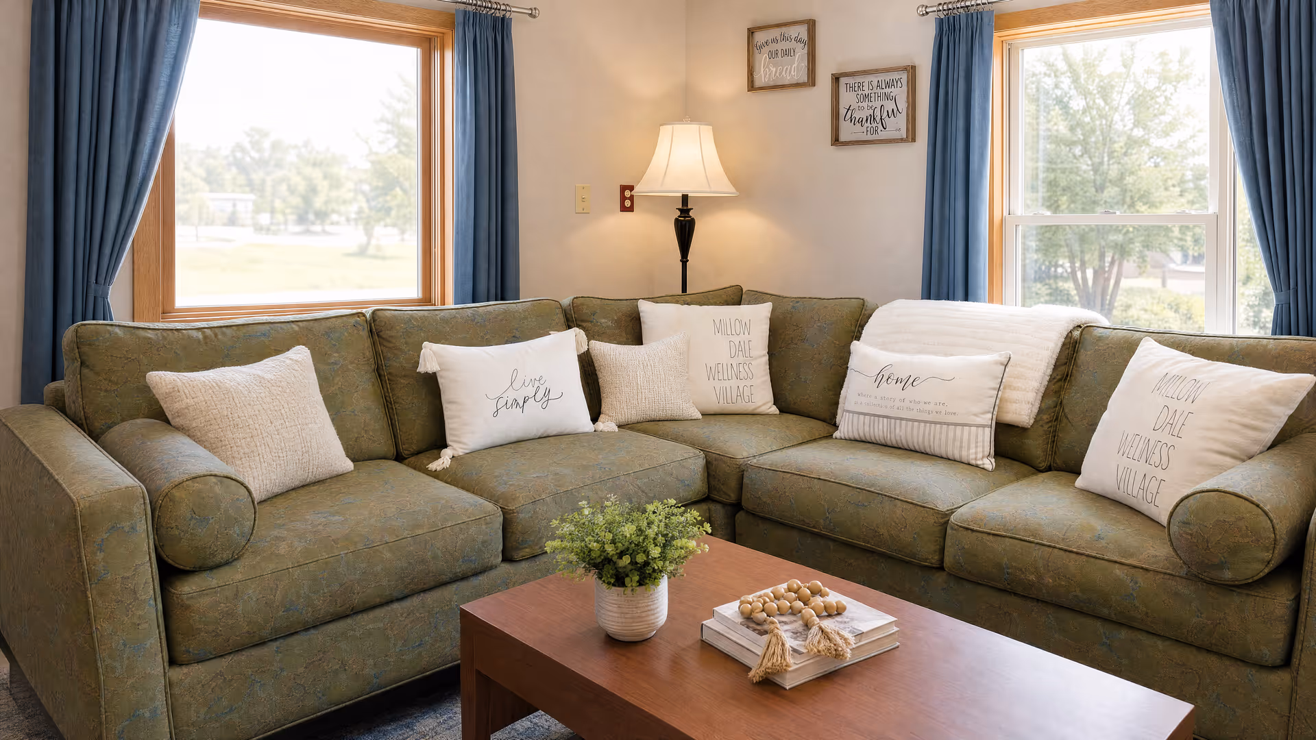 Cozy living room scene with green sectional, pillows, lamp, and blue-curtained windows.
