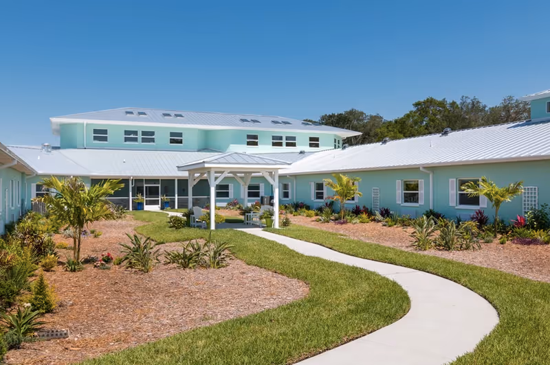 Exterior courtyard and entrance of a light-blue assisted living building with a curved walkway, landscaping, and a small white gazebo.