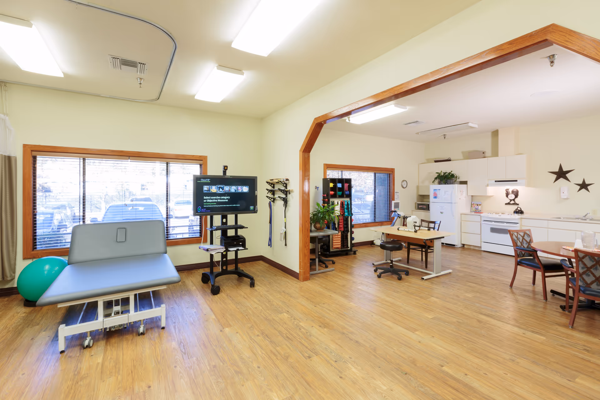 A spacious room with wooden flooring featuring a physical therapy or medical examination table, a large green exercise ball, a TV on a mobile stand displaying exercise options, and a rack with various exercise equipment. The room also includes a kitchen area with white cabinets, a refrigerator, stove, and a dining table with chairs. Large windows with wooden frames allow natural light to enter the room.