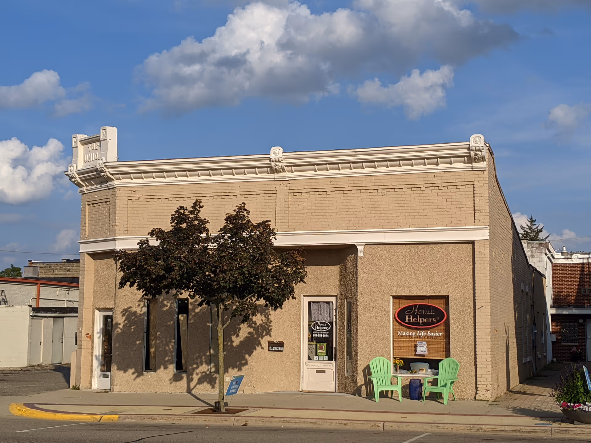 Beige brick building facade with a tree, door, window sign, and two green chairs.