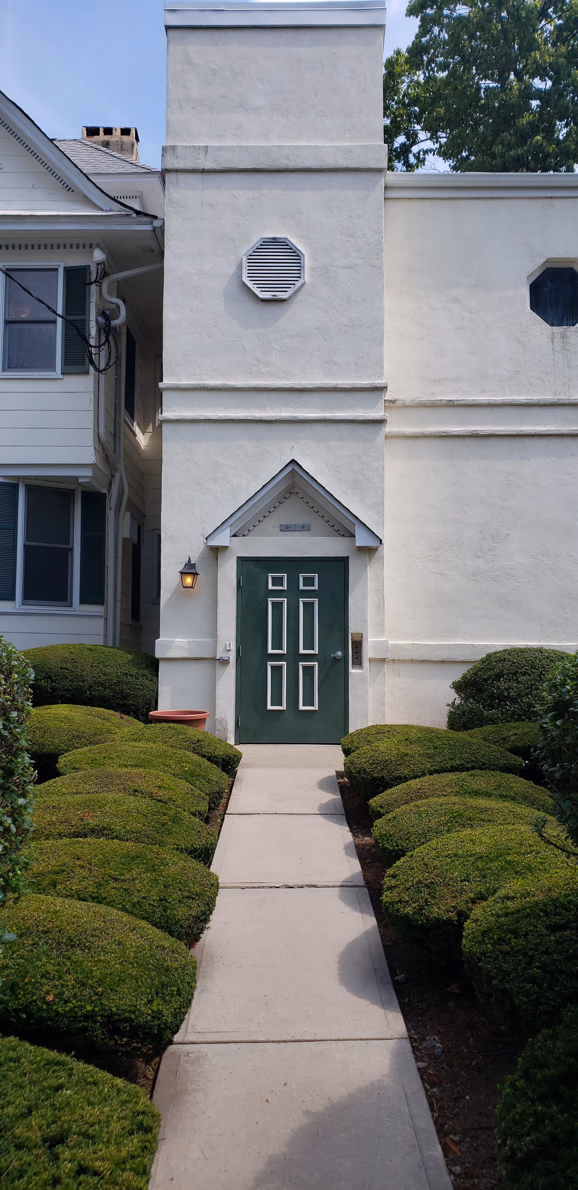 White building front with green door and neatly trimmed hedges along a concrete walkway.