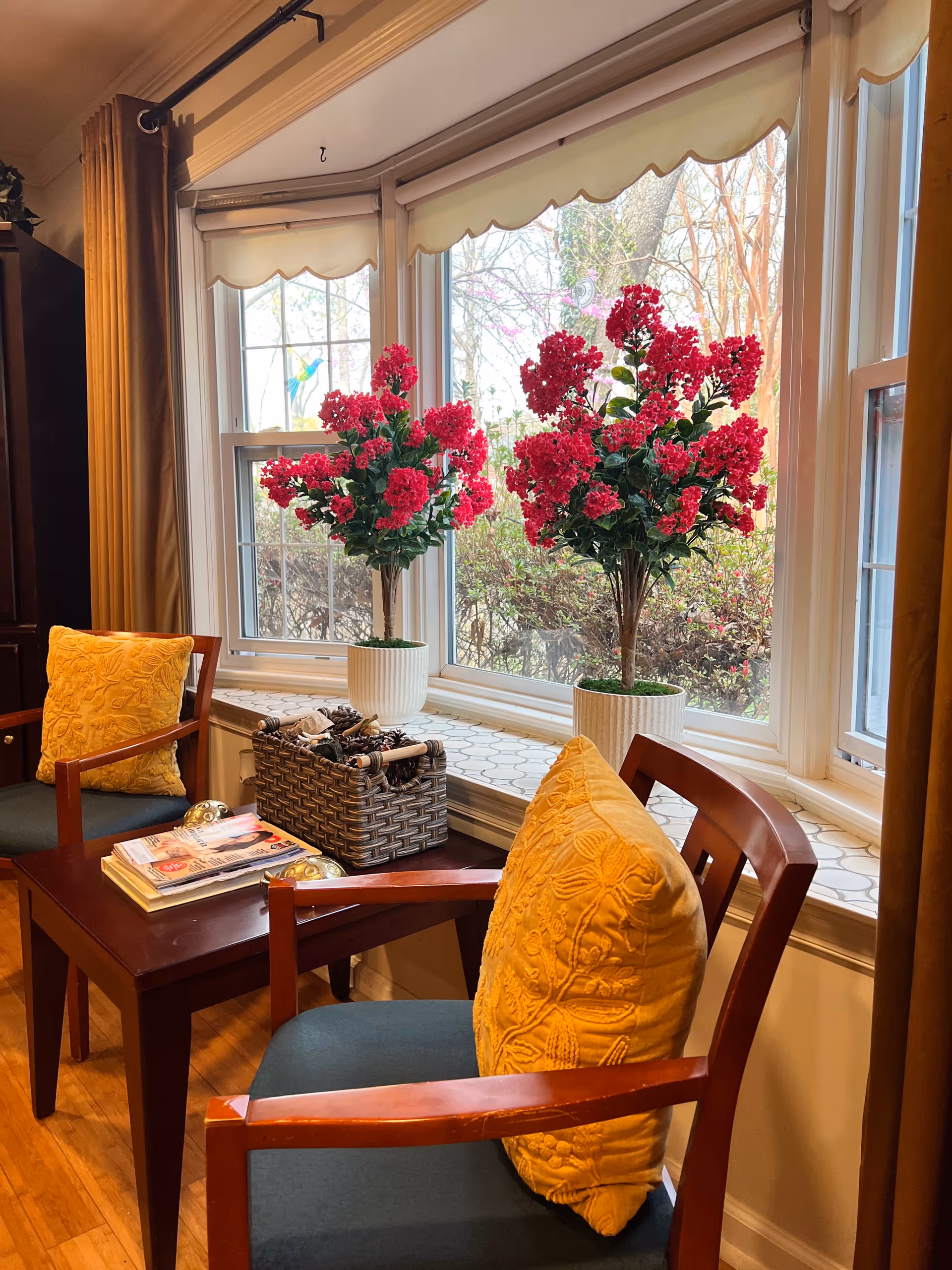 A cozy sitting area by large windows with red flowering plants, magazines, and yellow cushions.