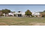 Exterior view of a single-story senior living facility with a flag, trees, and driveway.