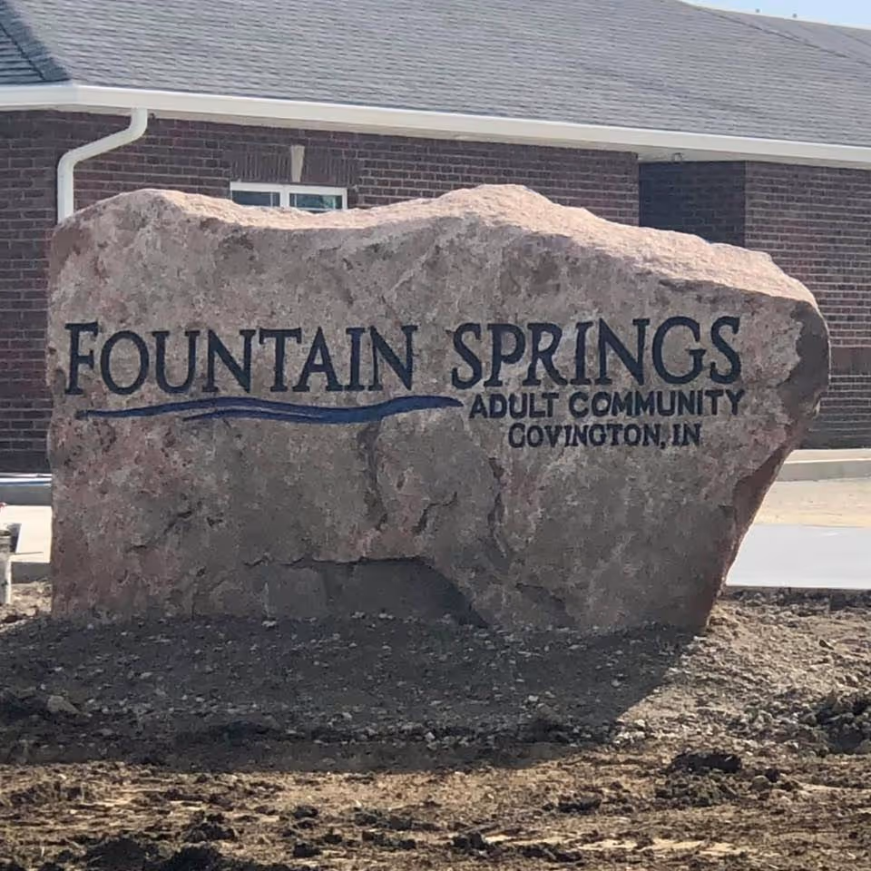 Large rock sign reads Fountain Springs Adult Community Covington, IN, in front of brick building.