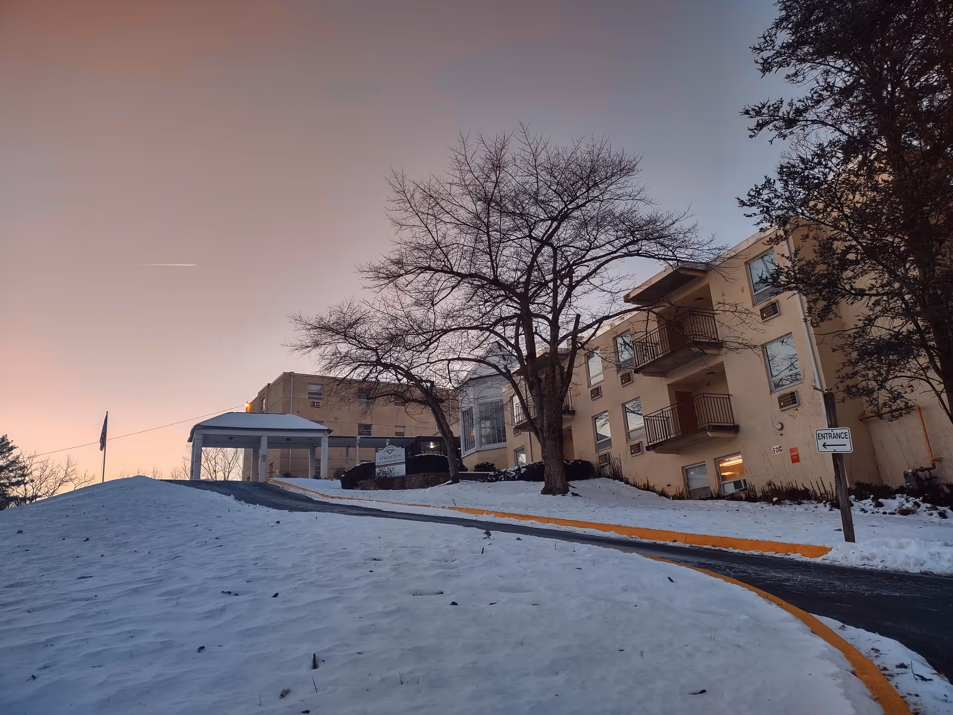 Snow covered slope leads to a beige senior living building with trees and an entrance sign.