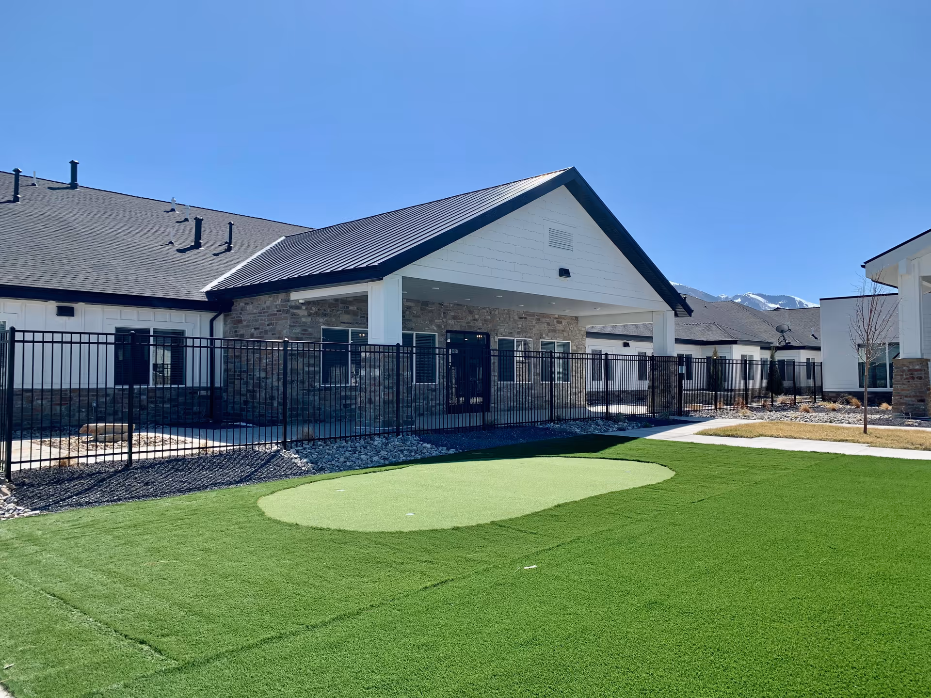 Exterior view of a single-story assisted living building with a fenced courtyard and a small artificial turf putting green.