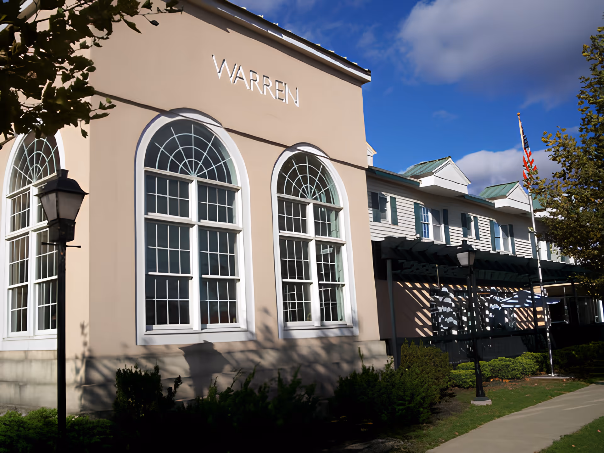 Exterior view of a senior living facility building with large arched windows and the word 'WARREN' displayed on the wall. The building has a beige facade with white trim, a green roof, and an American flag on a flagpole. There are trees, bushes, and a sidewalk in front of the building under a blue sky with some clouds.